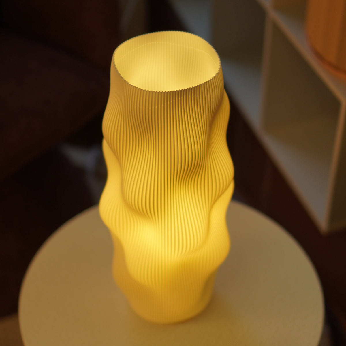 A glowing yellow sculptural lamp with a ribbed, wavy surface sits on a round table in a dimly lit interior setting.