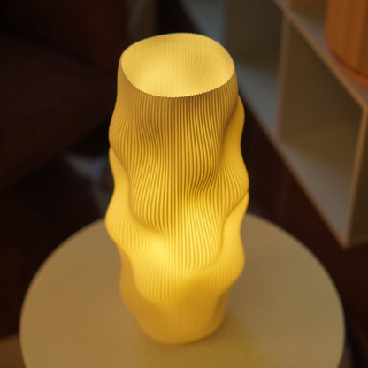 A glowing yellow sculptural lamp with a ribbed, wavy surface sits on a round table in a dimly lit interior setting.