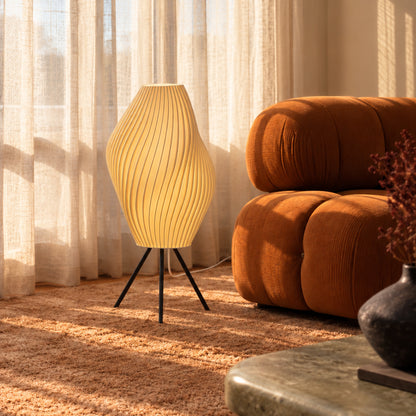 Crème Pleat Floor Lamp with a warm glow beside a rust-coloured lounge chair and sheer curtains.
