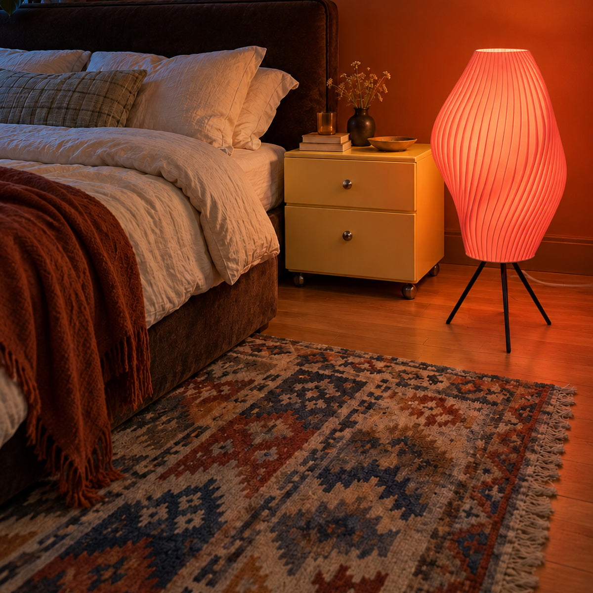 Flamingo pink Pleat Floor Lamp illuminated on its black tripod beside a bed in a warm-toned bedroom.