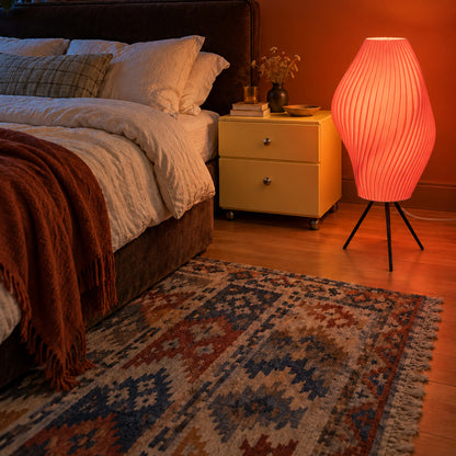 Flamingo pink Pleat Floor Lamp illuminated on its black tripod beside a bed in a warm-toned bedroom.